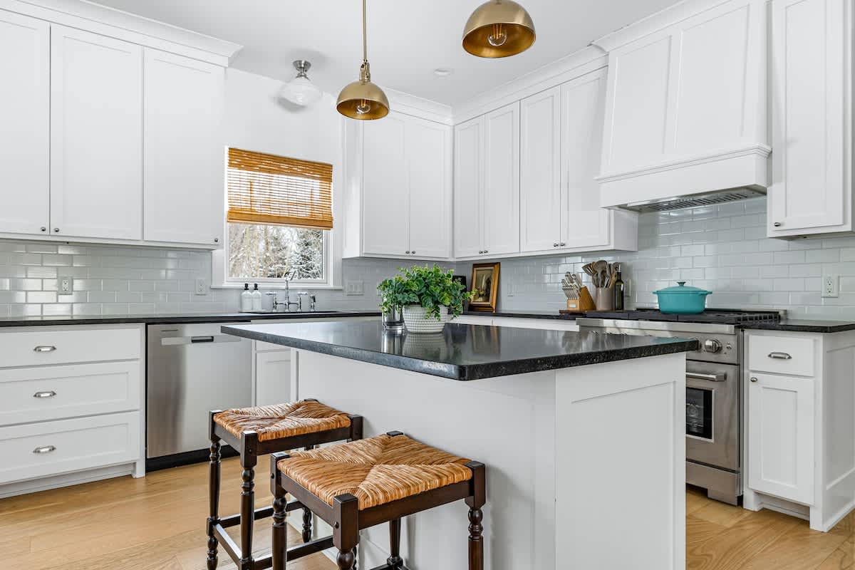 A well-lit kitchen showcases white shaker cabinets, dark countertops, and a white subway tile backsplash. A window with a bamboo blind is above the sink, and a stainless steel dishwasher is integrated next to the sink.