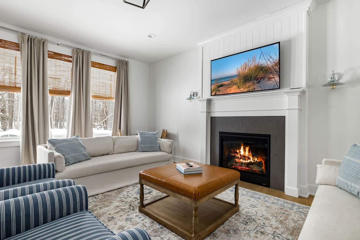 A bright living room with white walls and light wood floors features a cozy fireplace with a mounted TV above it. A white sofa and striped armchairs are arranged around a leather ottoman, with large windows covered by bamboo blinds and drapes.