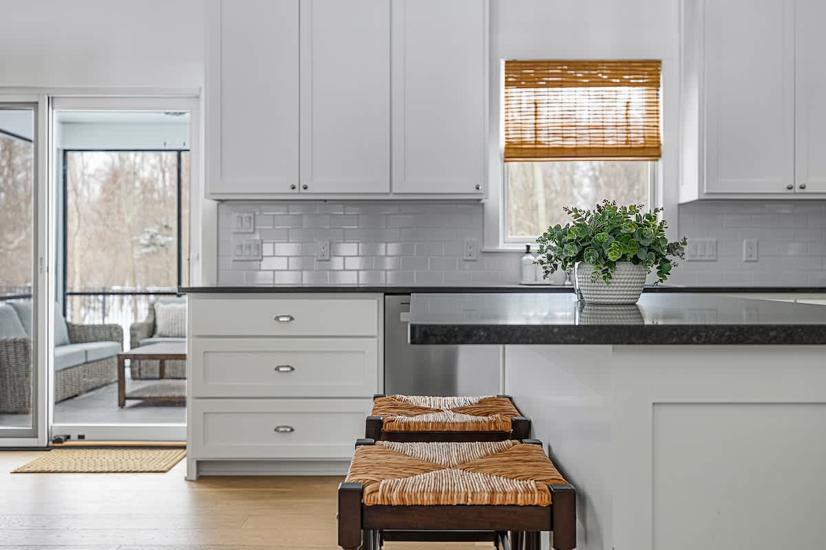 A modern kitchen features white shaker cabinets and dark countertops, with a white subway tile backsplash. Two woven-seat bar stools are pulled up to the kitchen island, and a sliding glass door leads to an outdoor screened porch.