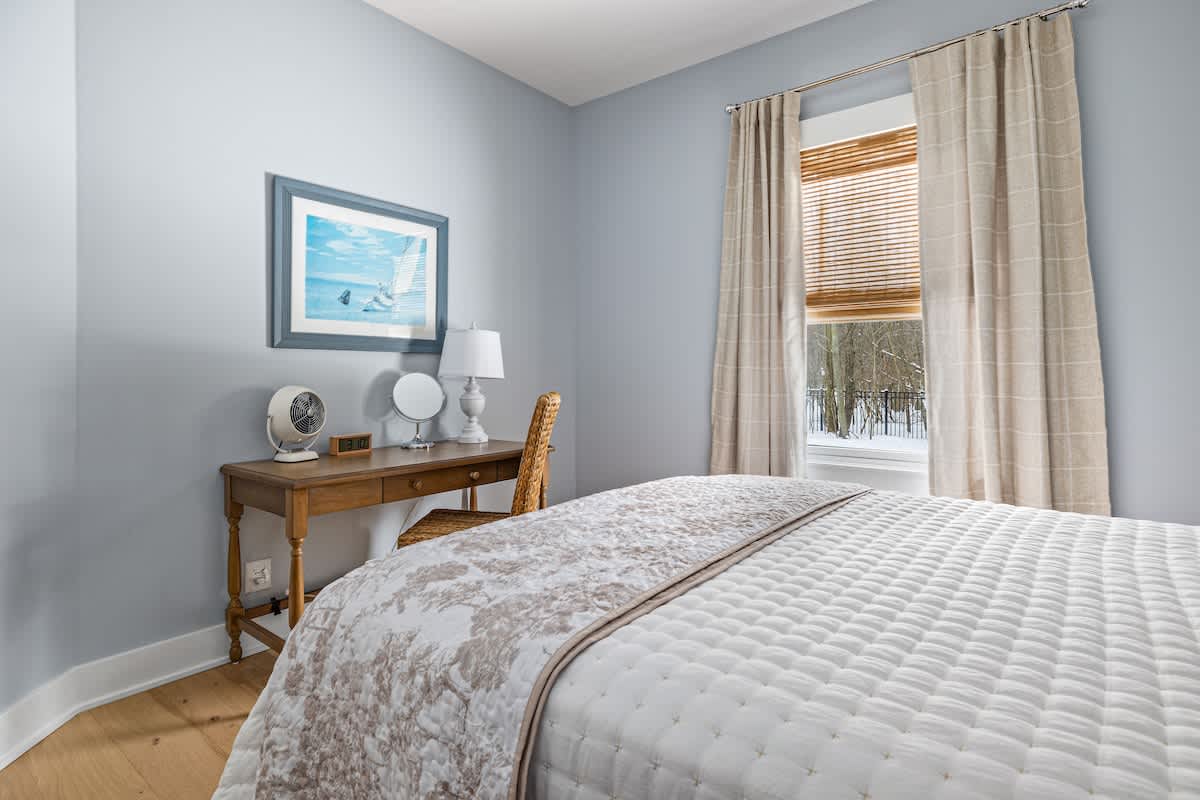 View of a light blue bedroom featuring a wooden desk with a lamp and mirror, and a white quilted bed with a patterned throw. The window is adorned with bamboo blinds and light-colored curtains.