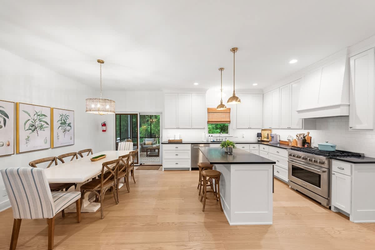 An open-concept kitchen and dining area features white shaker cabinets, dark countertops, and stainless steel appliances. A long white dining table with wooden chairs and a striped armchair is illuminated by a crystal chandelier, while the kitchen island has brass pendant lights.