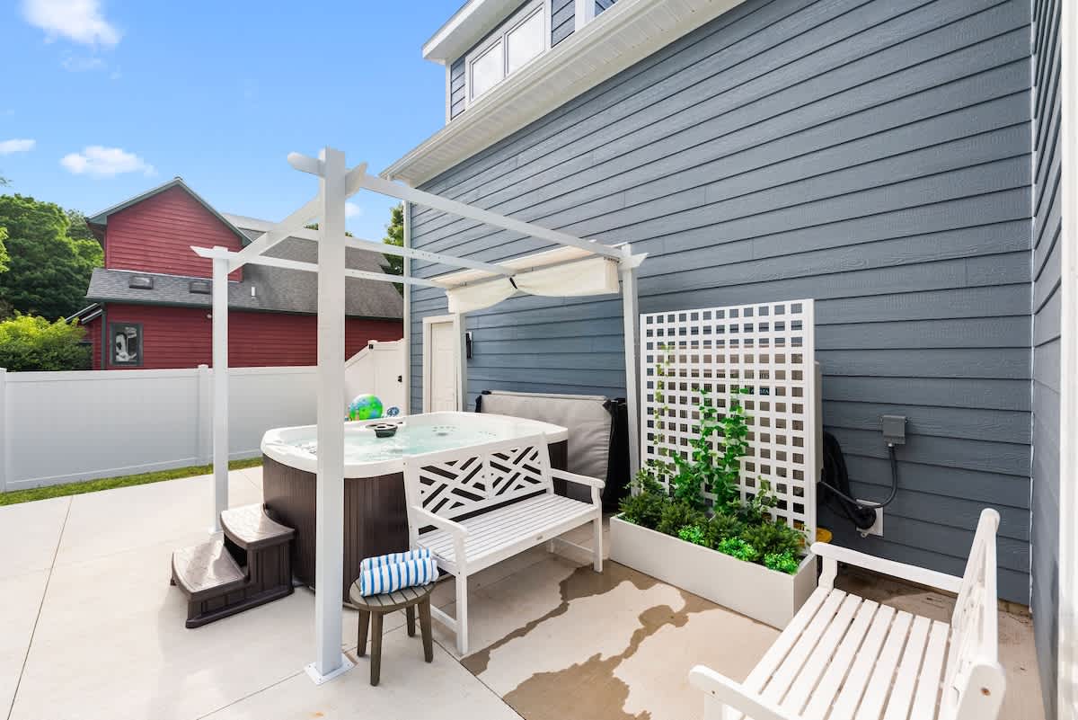 An outdoor patio area featuring a hot tub under a white pergola with a canopy, flanked by a white slatted bench and a decorative lattice screen with greenery.