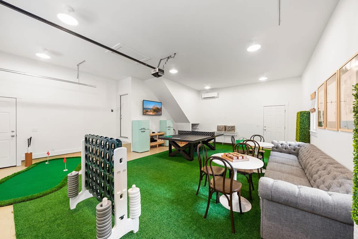 Expansive game room with a putting green, oversized Connect Four, and a ping pong table. A gray tufted sofa and small round tables provide seating, all set on a green artificial turf floor.