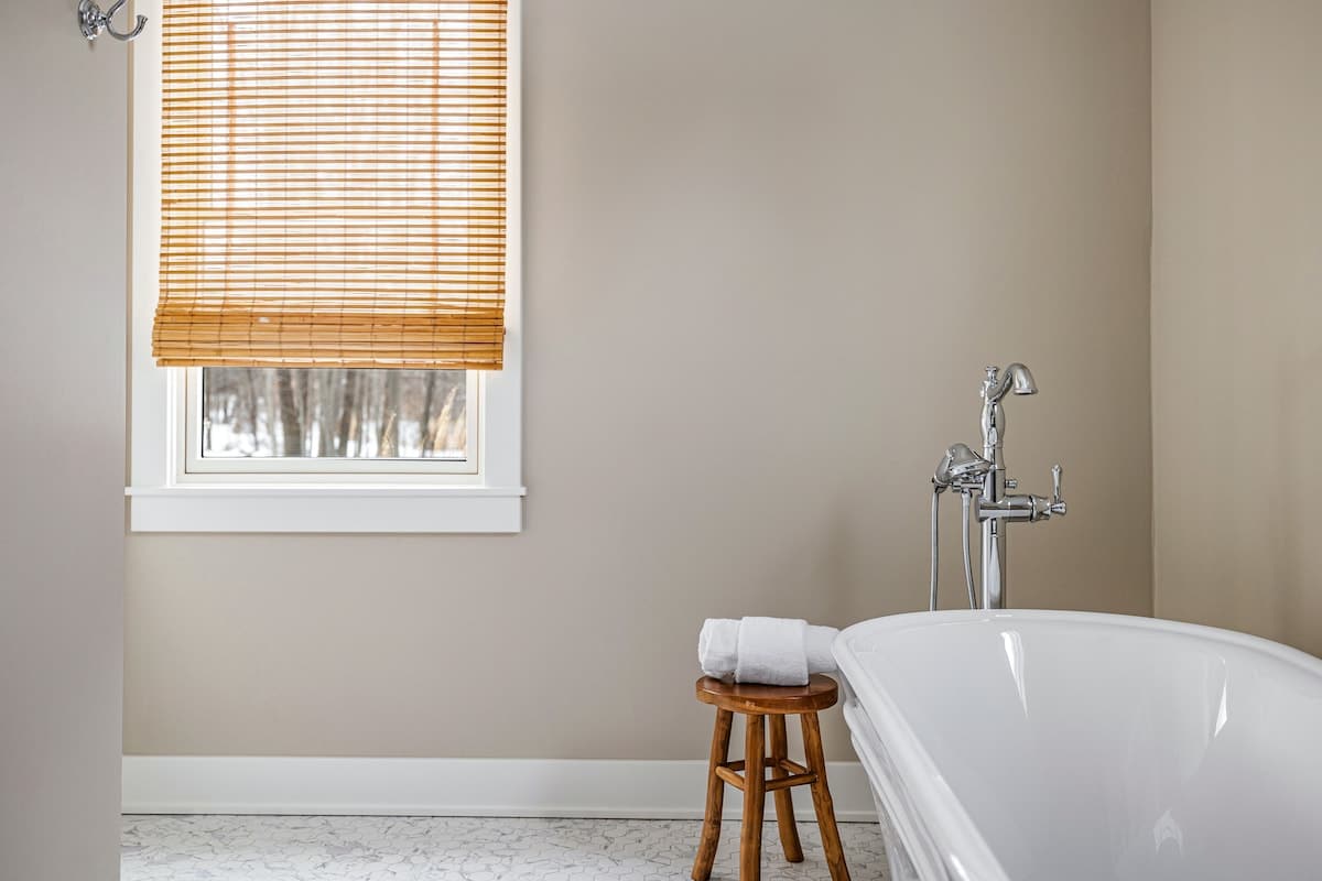 Close-up of the elegant freestanding white bathtub with a chrome floor-mounted faucet in bathroom-1, positioned next to a small wooden stool with white towels. A window with a bamboo blind offers natural light, set against light beige walls.