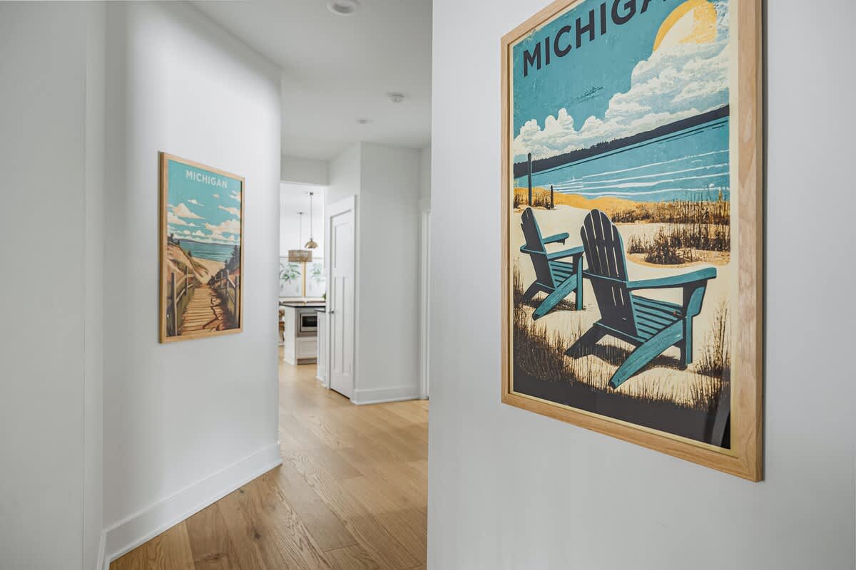 Bright hallway with light hardwood floors and white walls, adorned with two framed vintage-style travel posters depicting Michigan beach scenes. The hallway leads towards a kitchen area visible in the background.