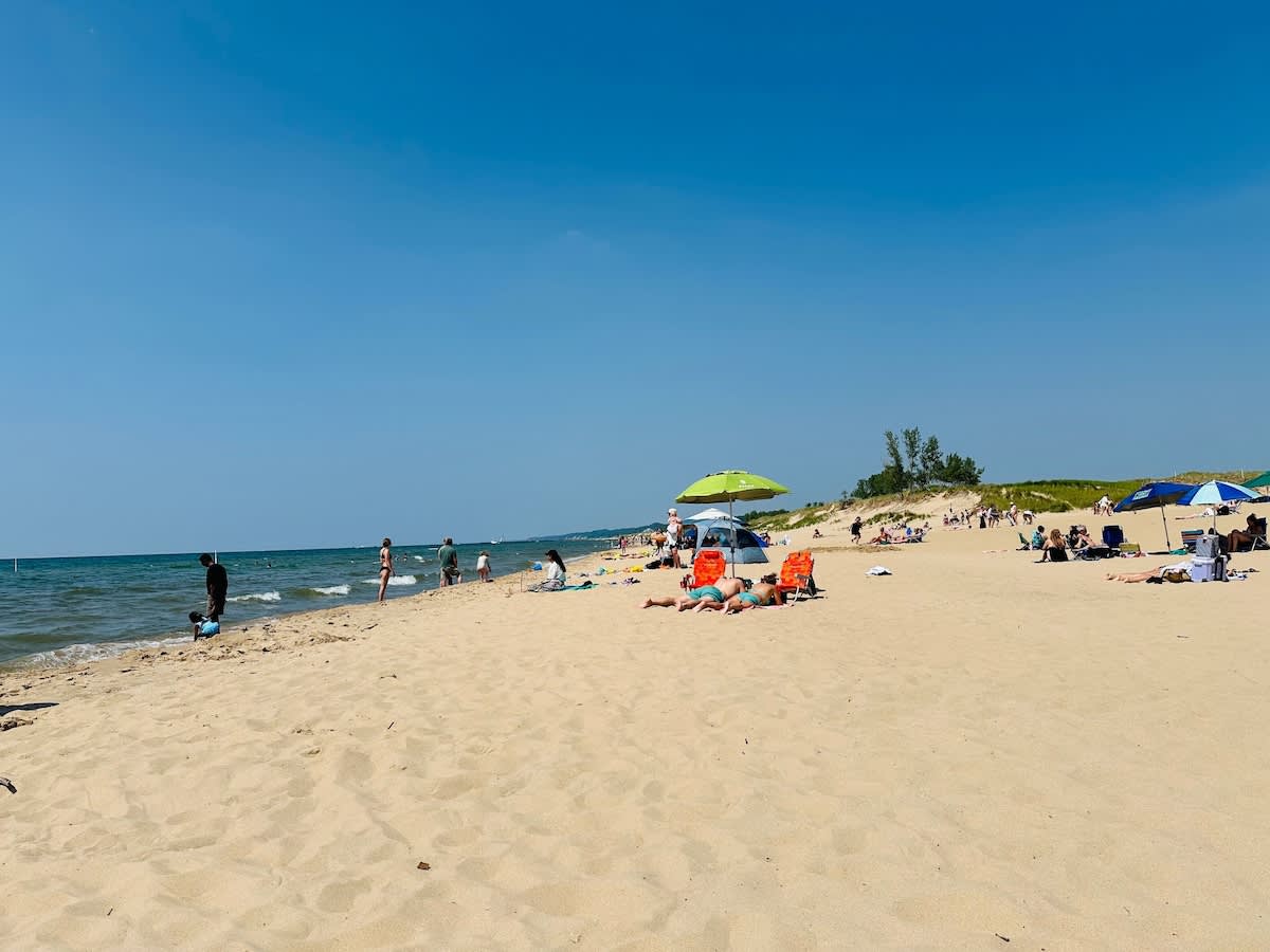A wide sandy beach stretches along a calm body of water under a clear blue sky, with several people relaxing on towels or under umbrellas. Gentle waves lap at the shore, and distant dunes are visible.