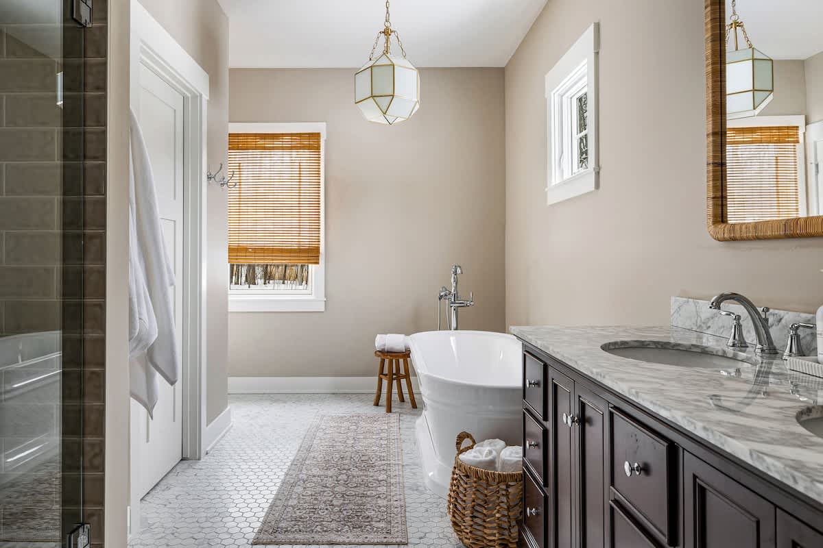Wide view of bathroom-1, showcasing the dark wood double vanity with a marble top and a large walk-in shower with subway tiles on the left. The freestanding tub is visible in the center, and a window with a bamboo blind adds warmth to the light beige space.