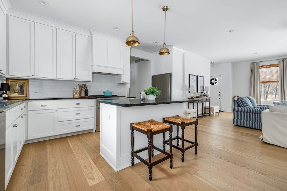 A bright kitchen features white shaker cabinets, dark countertops, and a white subway tile backsplash. A kitchen island with two woven-seat bar stools is illuminated by brass pendant lights, and a stainless steel refrigerator is visible in the background.