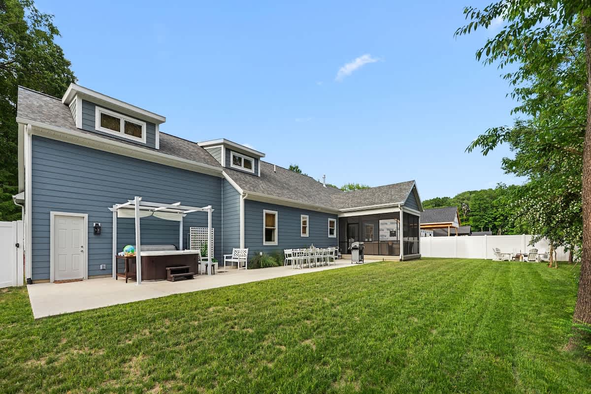 A side view of the blue-sided house with a large grassy backyard, a concrete patio with outdoor dining and a hot tub under a pergola, and a white privacy fence.