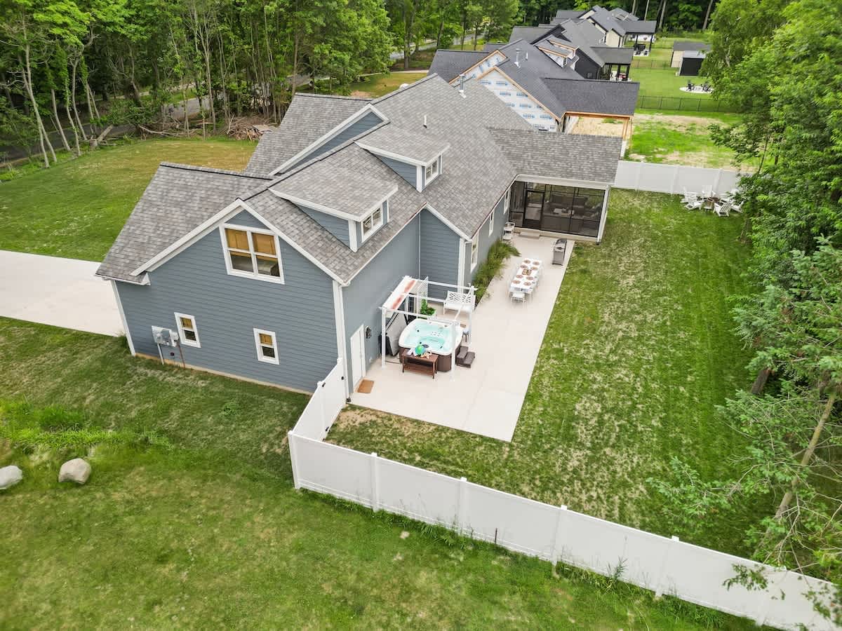 An aerial view of a blue-sided house with a gray roof, showing a large backyard with a white fence, a patio with a hot tub, and a screened-in porch.