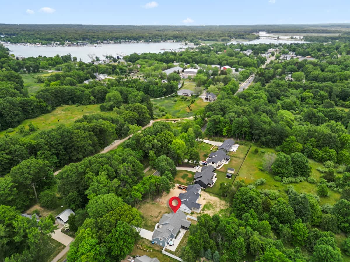 An aerial view shows a residential area surrounded by dense green trees, with a large body of water (likely a lake or river) in the background. A red pin highlights the property, which is part of a cluster of modern homes.
