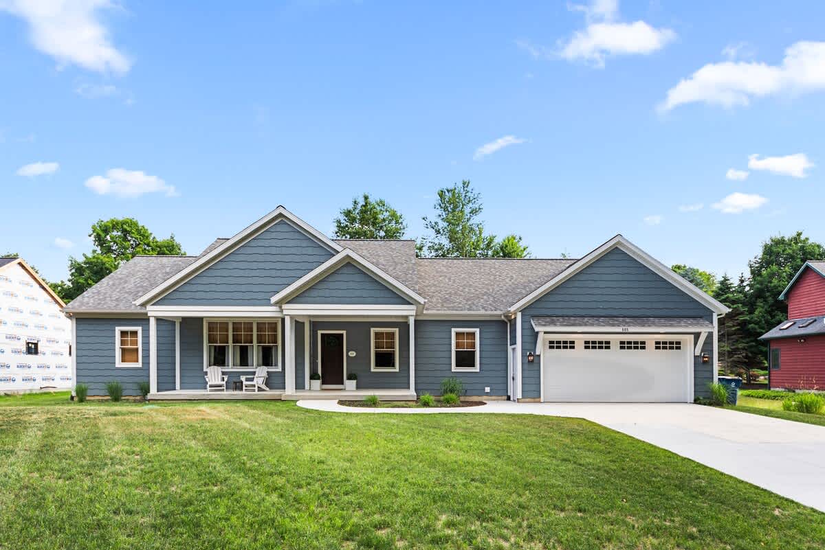 A modern ranch-style home with blue siding and white trim features a covered front porch with two white Adirondack chairs. A two-car garage with a white door is visible on the right, and the property is surrounded by a green lawn under a blue sky.