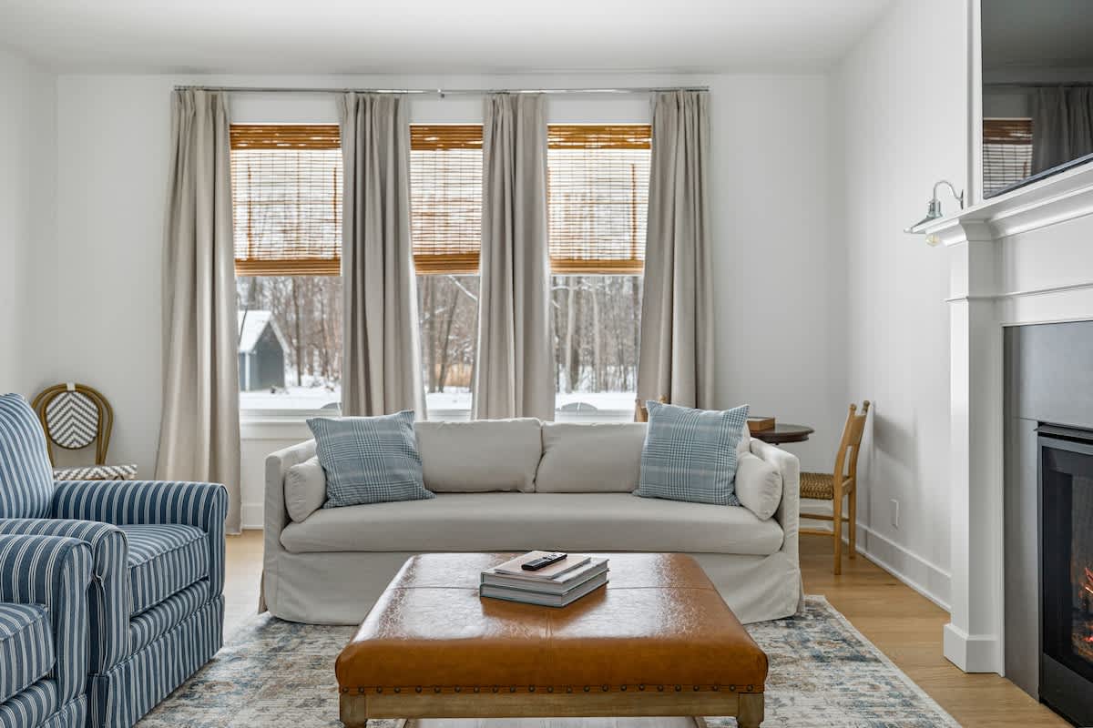 A bright living room features a light-colored sofa with blue plaid pillows, a striped armchair, and a large brown leather ottoman serving as a coffee table. Three windows with bamboo blinds and light drapes overlook a snowy outdoor scene.