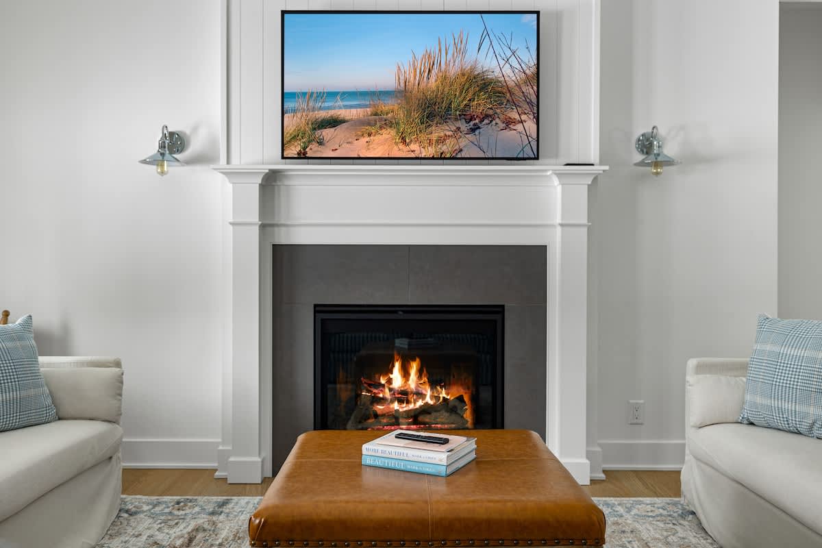 A cozy living room centers around a white fireplace with a large flat-screen TV mounted above, displaying a beach scene. A warm fire glows in the hearth, and a brown leather ottoman with books sits on an area rug in front.