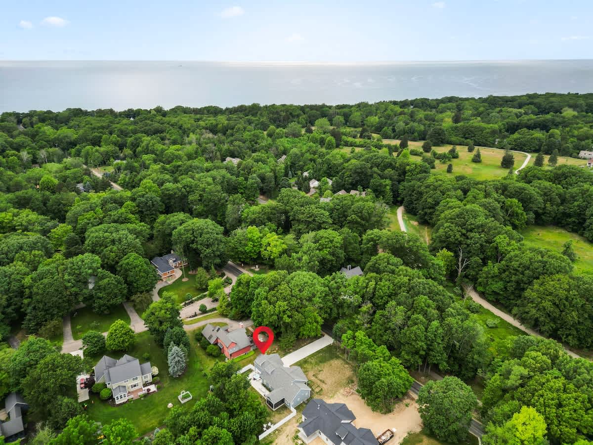 A high aerial view displays the property (marked with a red pin) surrounded by dense green forests, with a large body of water, likely a lake or ocean, visible in the distance under a clear sky.