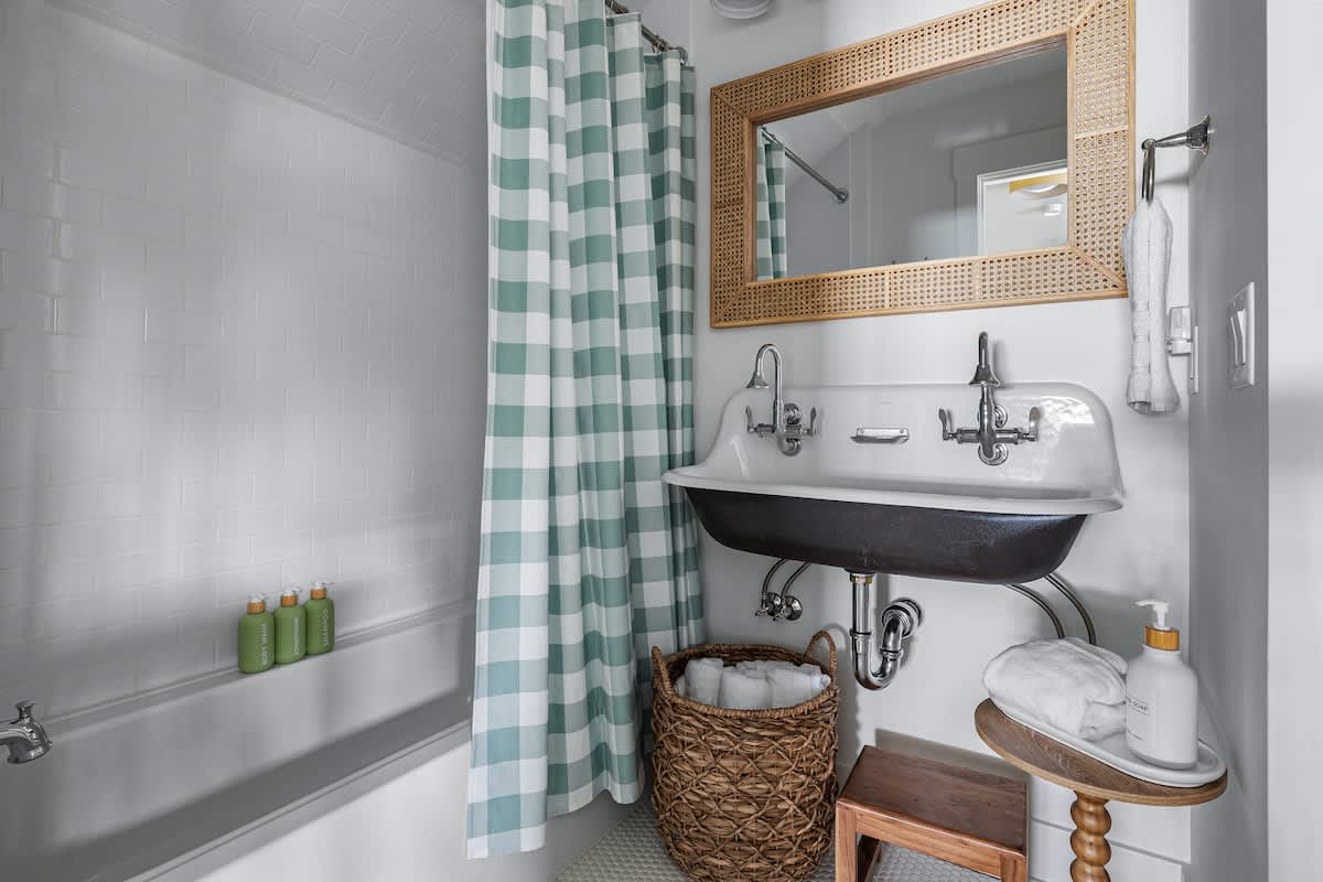 A bright bathroom with white subway tile walls, a bathtub with a green and white checkered shower curtain, and a vintage-style double basin sink with a rattan-framed mirror.