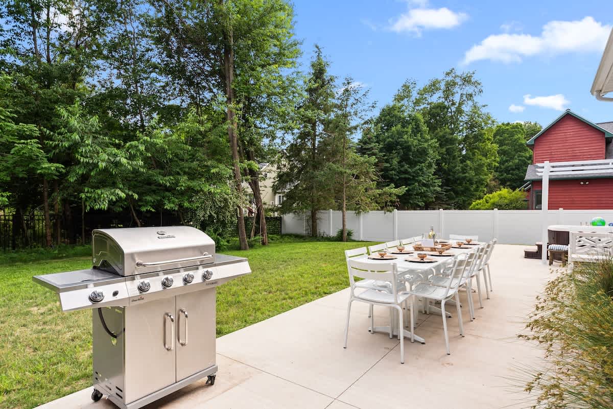 A spacious outdoor patio with a stainless steel BBQ grill and a long white dining table with chairs, set on a concrete slab next to a grassy lawn and mature trees.