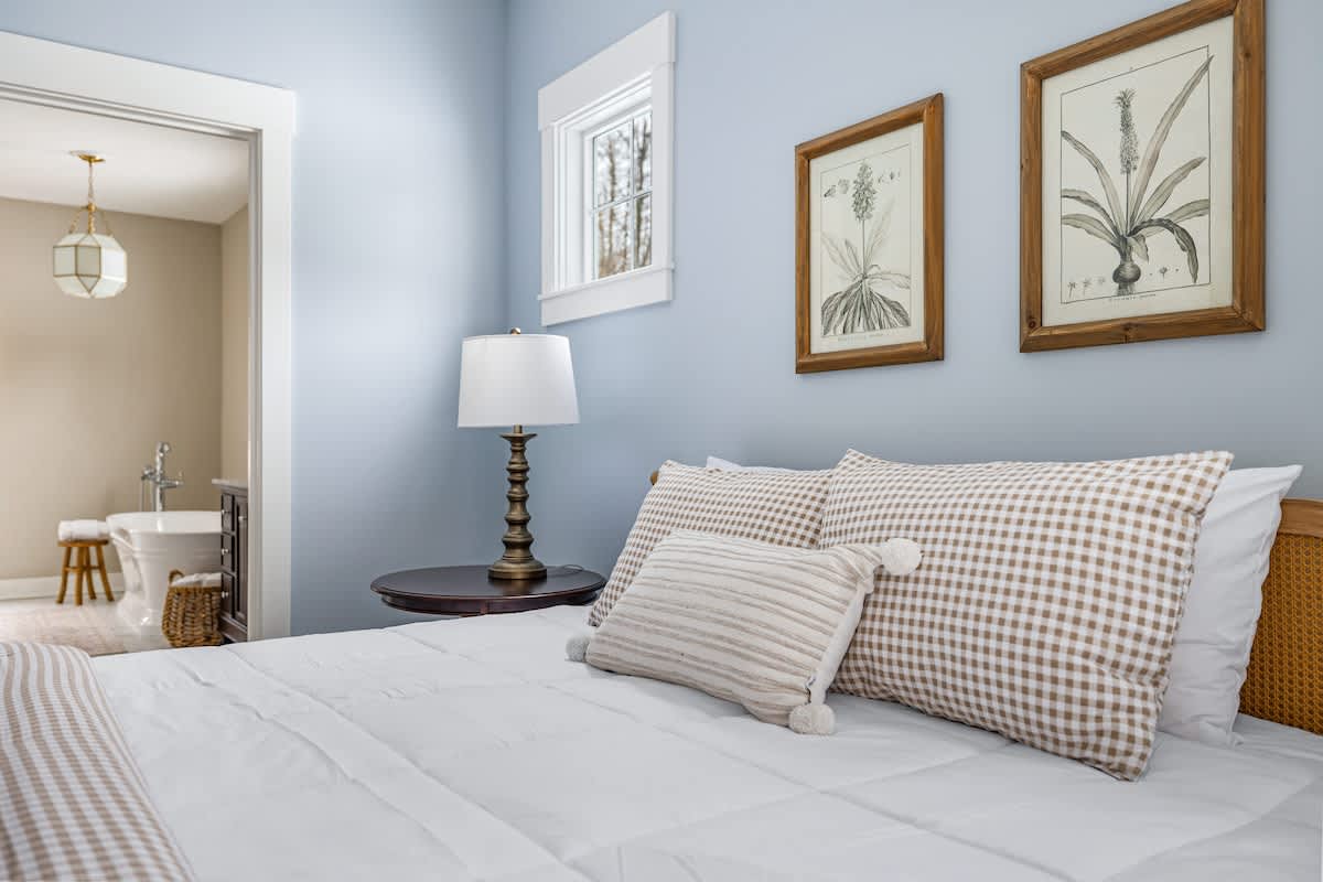 Close-up view of the bed in bedroom-1, showcasing the natural wood cane headboard, white duvet, and a mix of tan gingham and striped throw pillows. A dark wood nightstand with a textured lamp is visible to the left, and a window with white trim offers a glimpse of the outdoors.