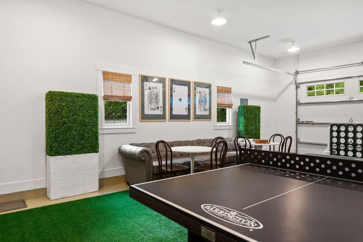 A bright game room, likely a converted garage, featuring a black ping pong table on a green artificial turf rug, a gray tufted sofa, and a small round table with chairs, all under large framed playing card artwork.