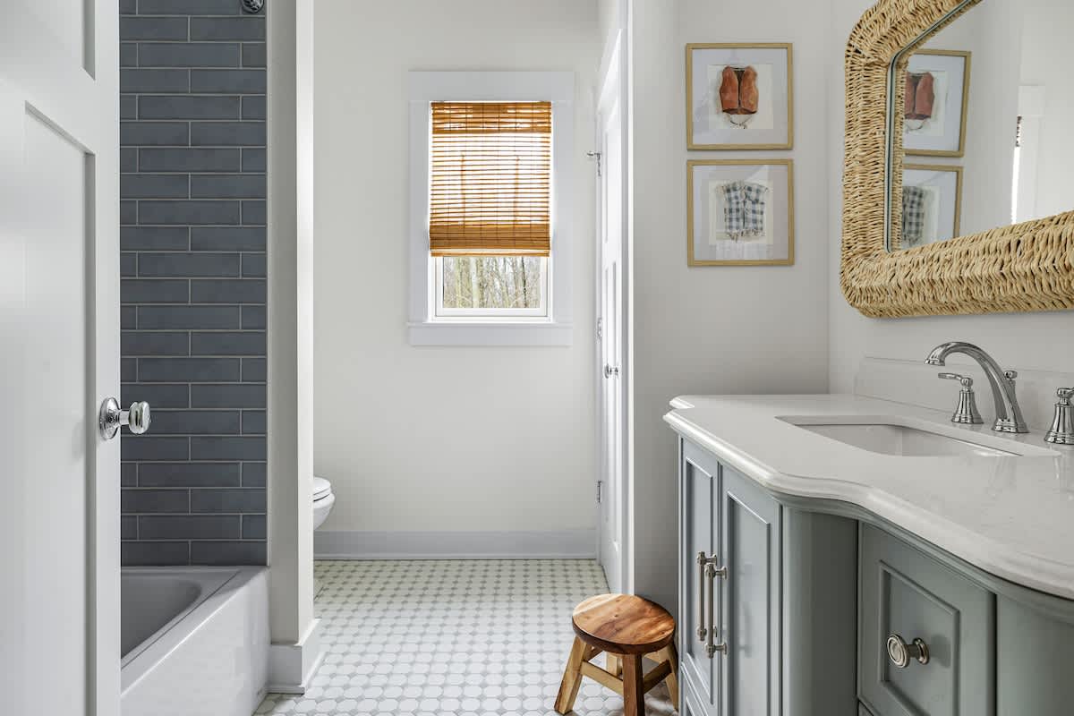 Bright bathroom with a gray vanity, white countertop, and a large woven mirror. A bathtub with dark gray subway tile walls is visible to the left, and a small wooden stool sits on the patterned floor.