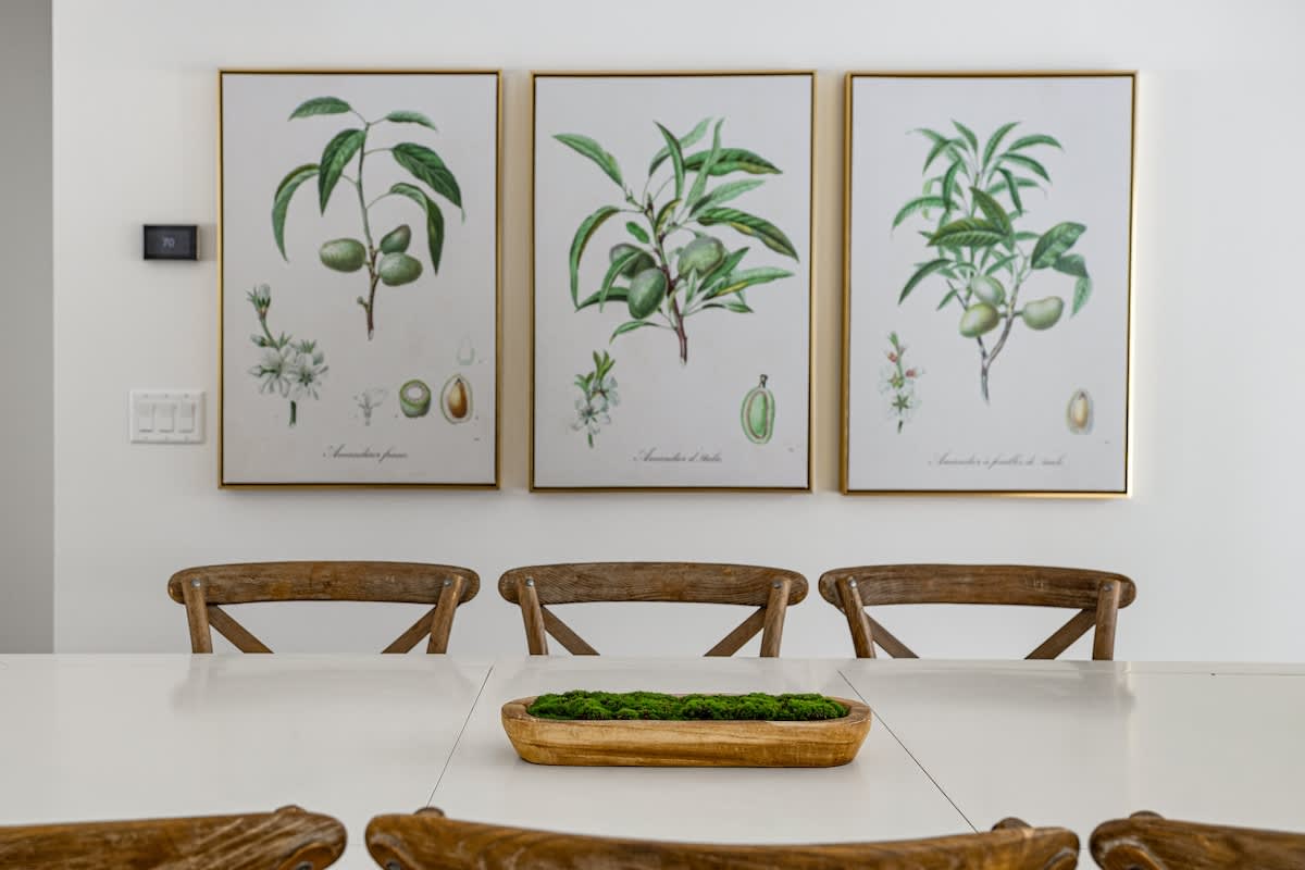 A close-up of the dining area shows a long white table with a wooden bowl centerpiece filled with moss. Three botanical prints in gold frames adorn the white wall above several wooden cross-back dining chairs.