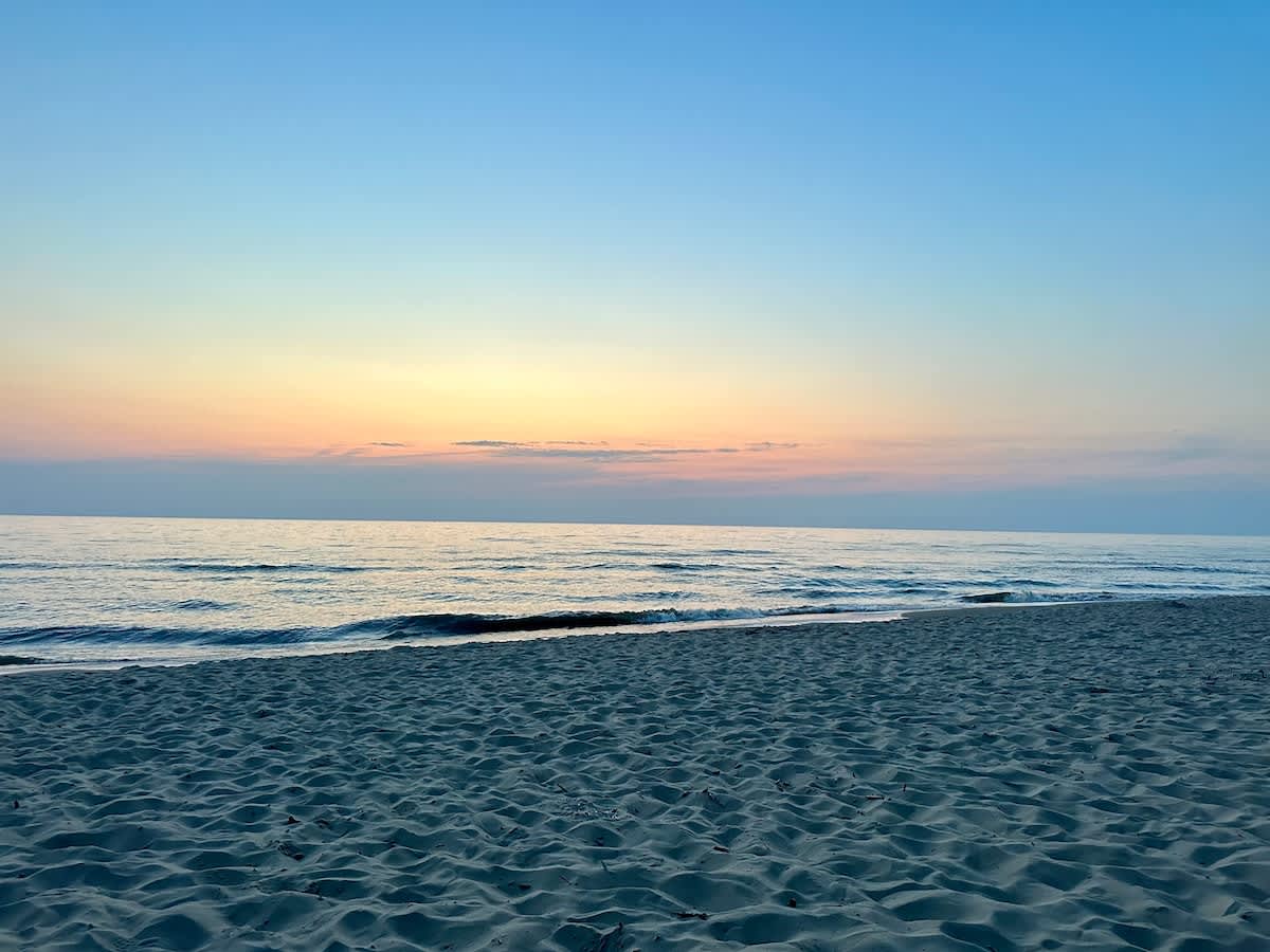 A serene beach scene at sunset, with soft waves gently rolling onto the sandy shore under a gradient sky of blue, pink, and orange hues. The vast ocean stretches to the horizon, meeting the colorful sky.