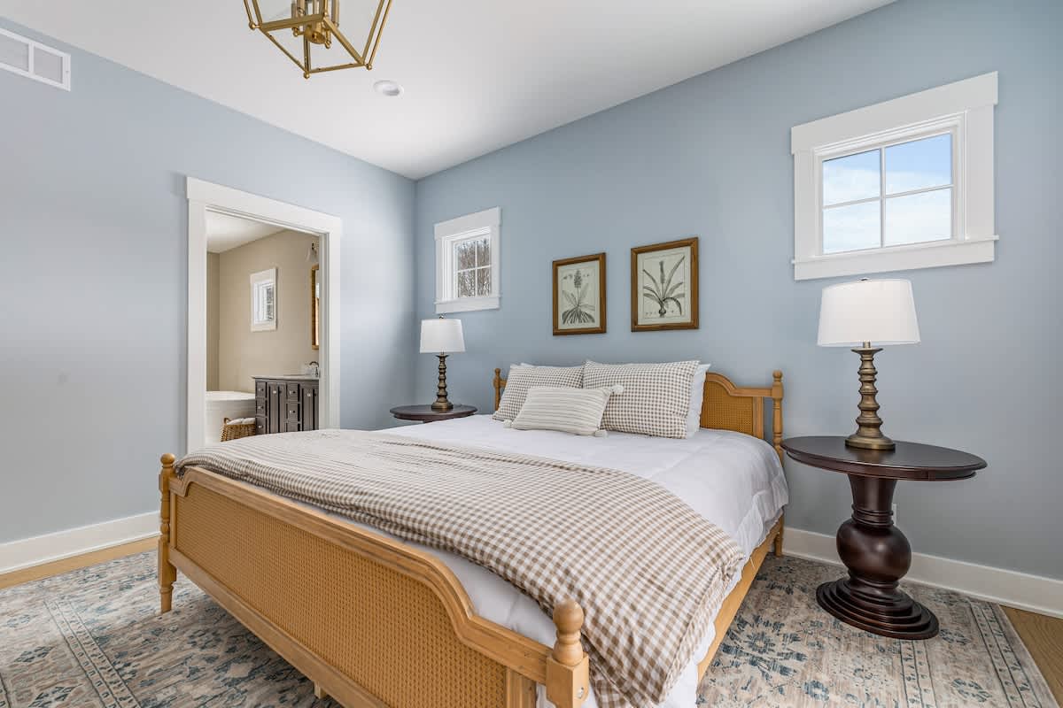 Serene bedroom with light blue walls and light wood flooring, featuring a natural wood cane bed frame dressed in white bedding with a tan gingham throw. Two dark wood pedestal nightstands flank the bed, each with a decorative lamp, and two botanical prints hang above the headboard.