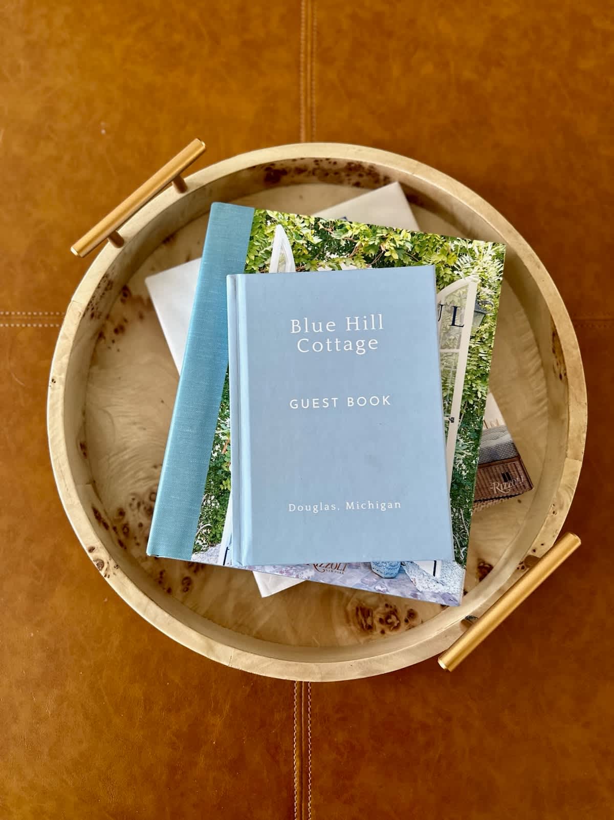A close-up shot of a light blue guest book titled "Blue Hill Cottage, Douglas, Michigan" resting on a wooden tray with gold handles. The tray is placed on a brown leather surface, likely an ottoman or table.