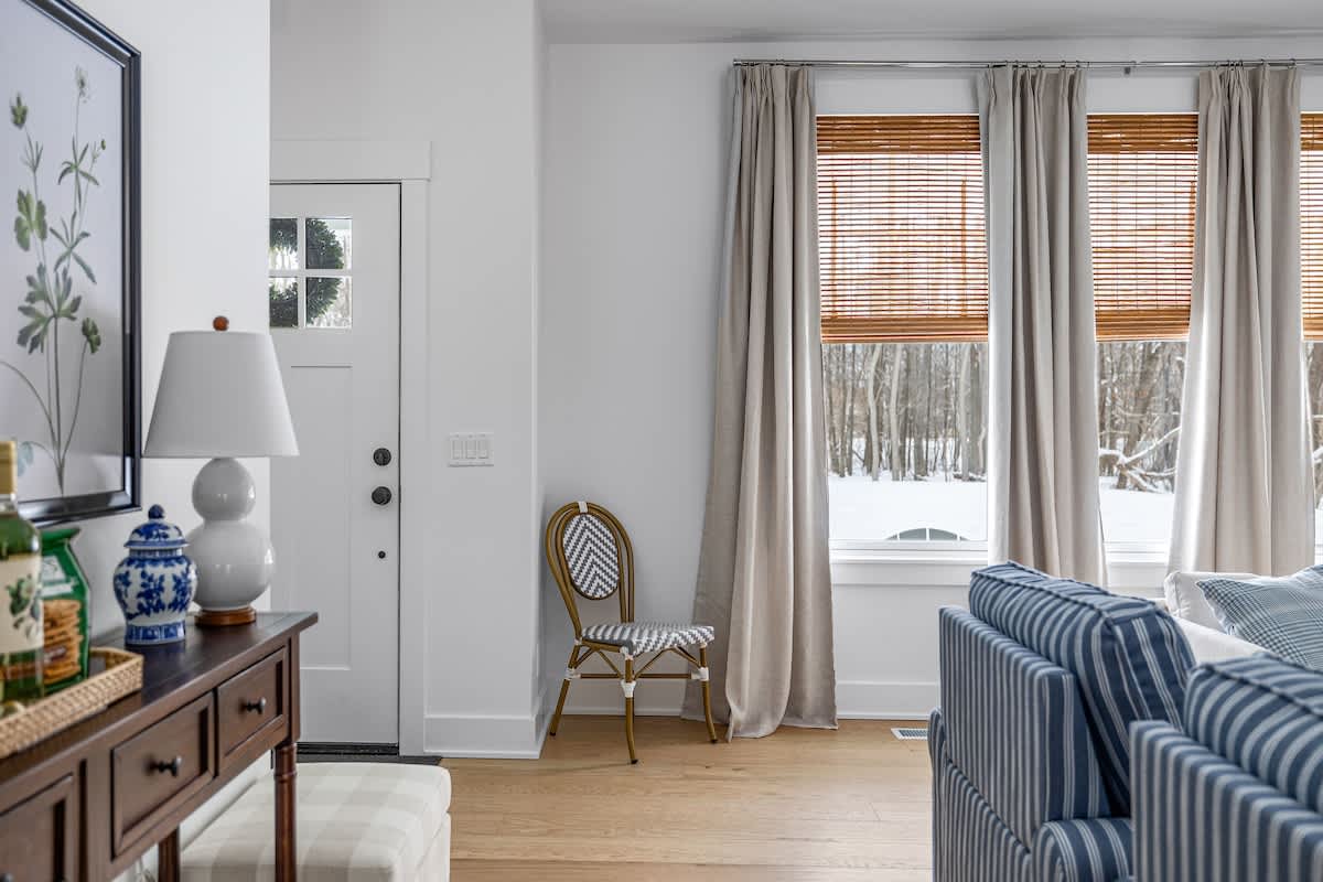 A bright entryway or corner of a living room with white walls and light wood floors, featuring a dark wood console table with decor. A white front door with a wreath is visible, and large windows with bamboo blinds and drapes look out onto a snowy landscape.