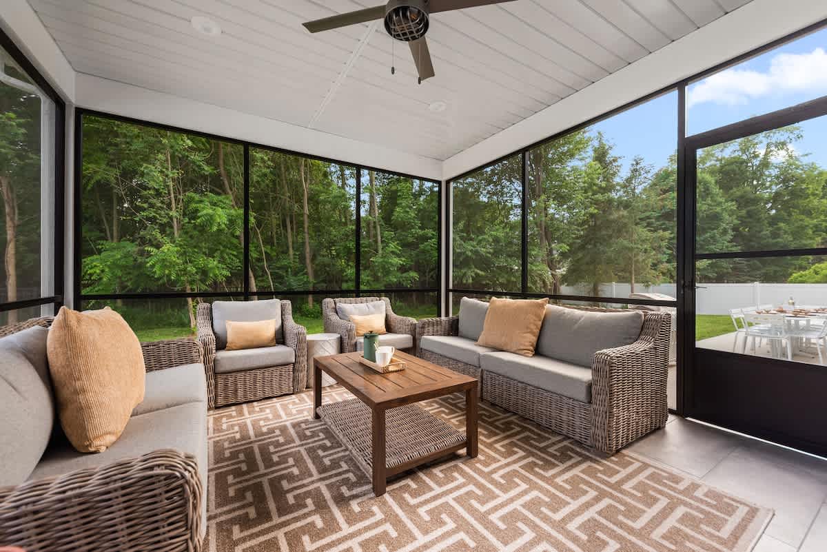 A spacious screened porch features comfortable wicker seating with light-colored cushions and throw pillows, arranged around a wooden coffee table on a patterned rug. The screens offer views of lush green trees outside.