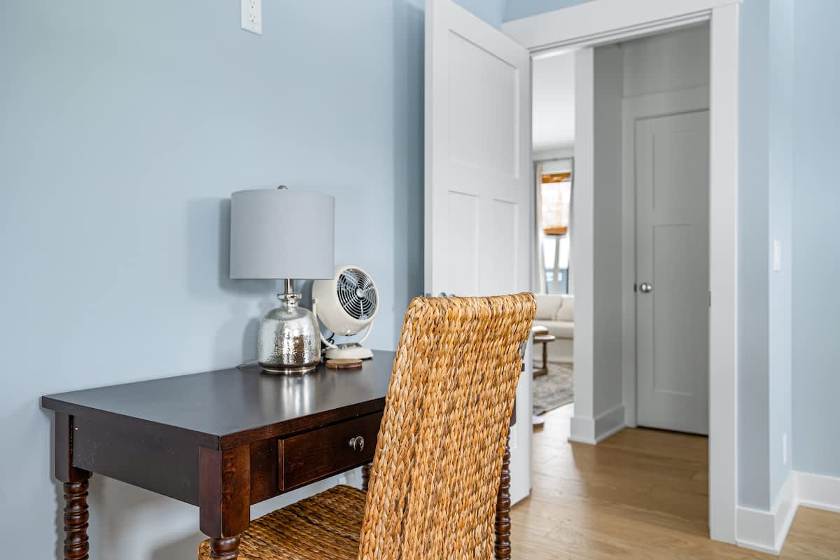 A small dark wood desk with turned legs and a woven wicker chair sits against a light blue wall in bedroom-1, featuring a silver lamp and a small white fan. A white door leads to an adjacent room, revealing a glimpse of a living area.