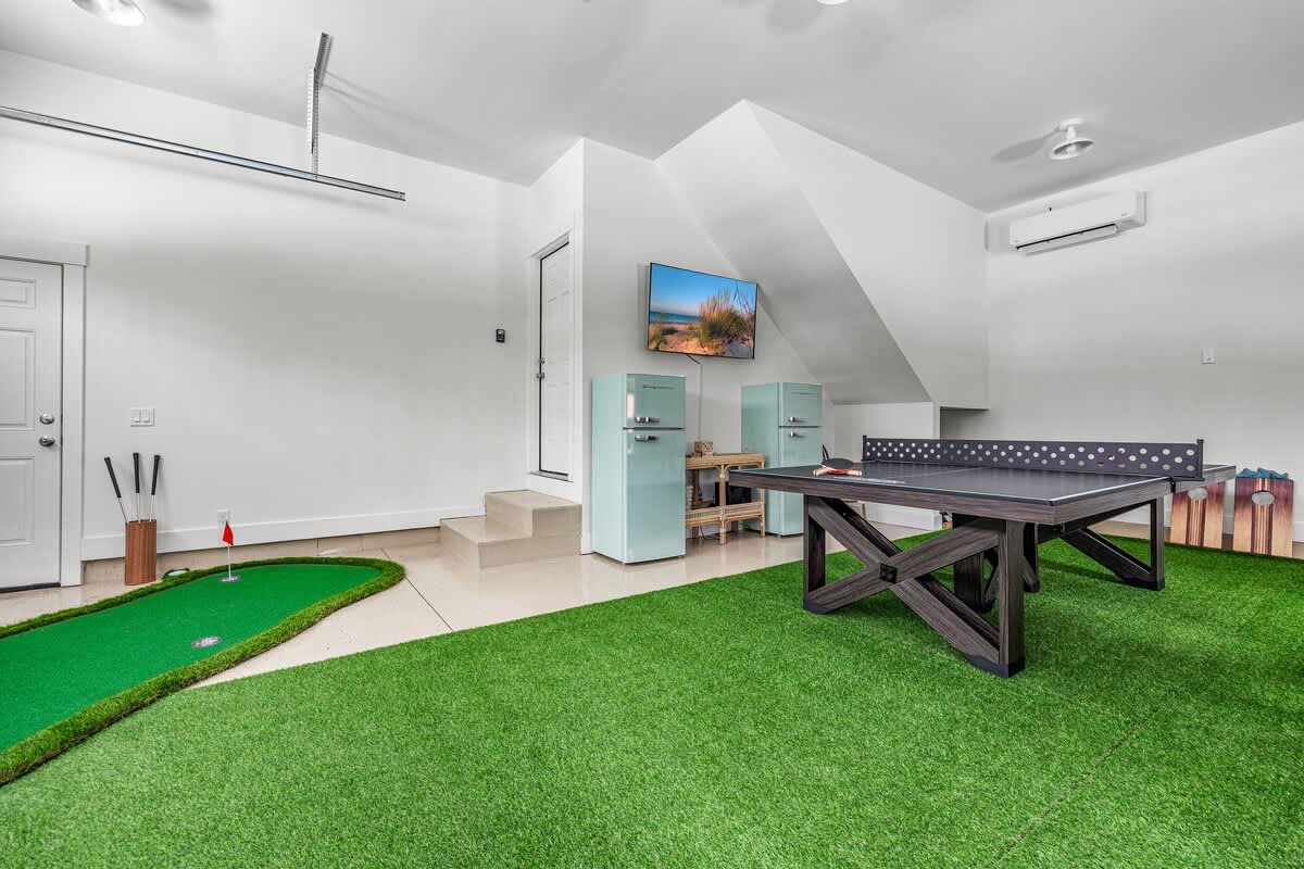 A spacious game room, set in a garage, with a putting green on artificial turf, a black ping pong table, two light blue retro-style mini-fridges, and a wall-mounted TV.