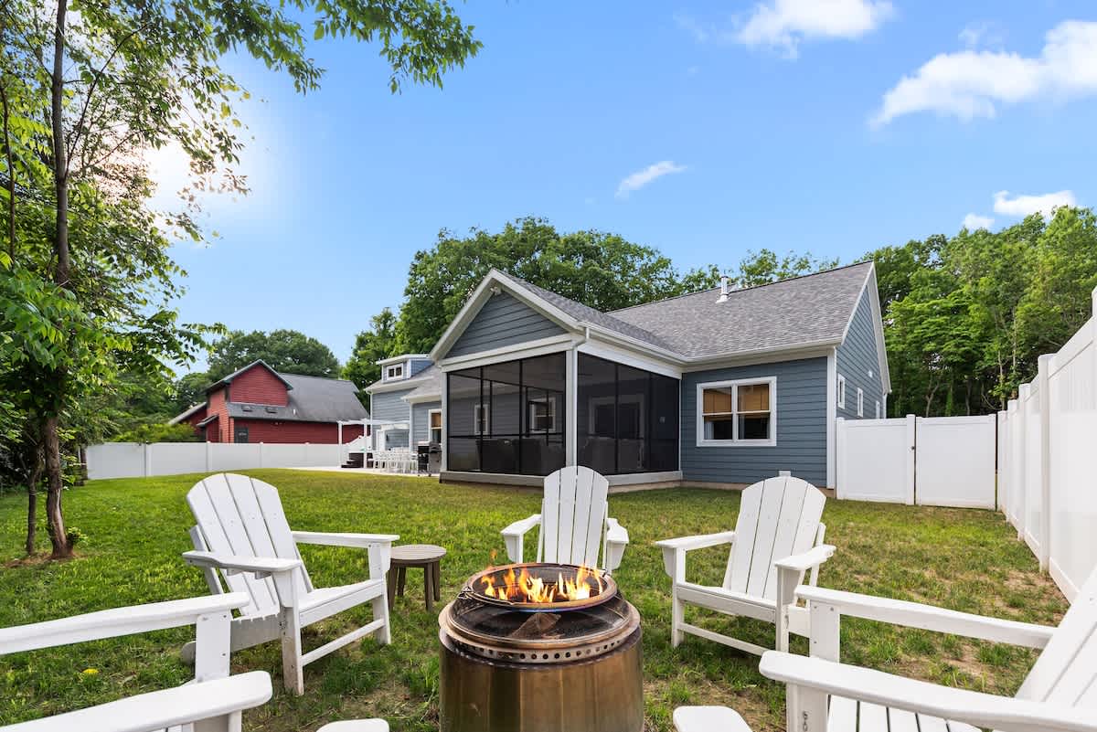 A sunny backyard featuring a circular fire pit surrounded by four white Adirondack chairs on a green lawn, with the blue-sided house and a screened porch in the background.