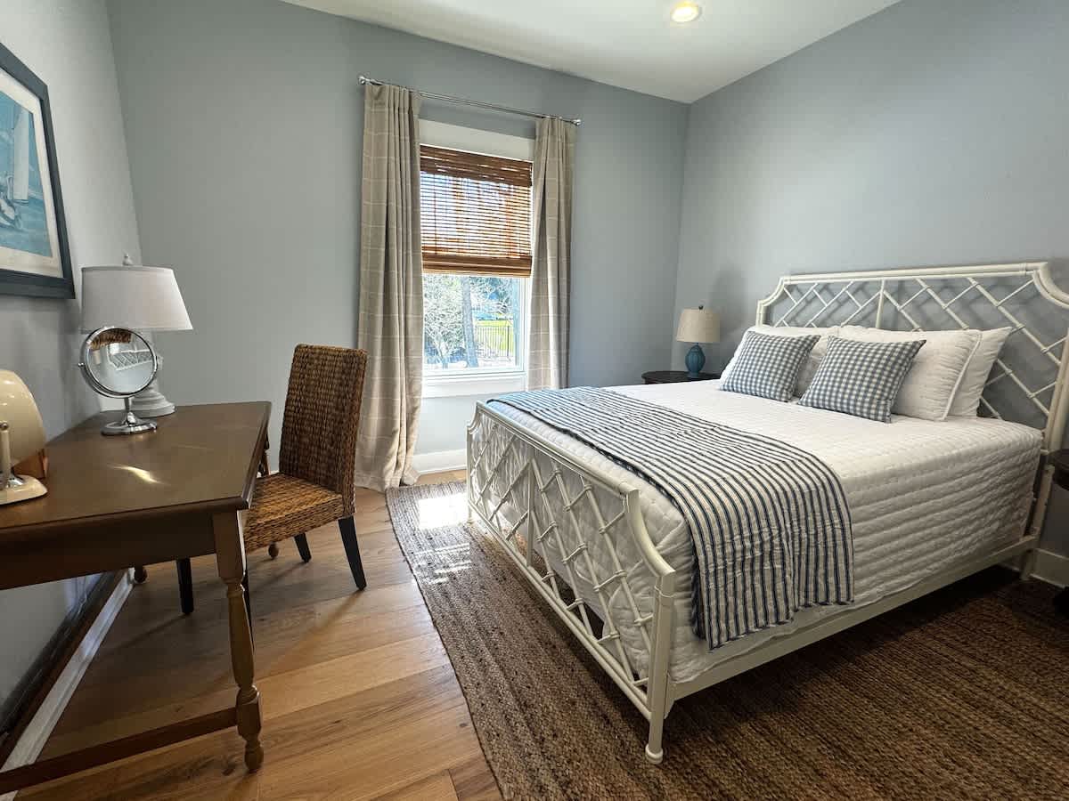 Serene bedroom with light blue walls and a white metal bed frame dressed in a striped throw. A wooden desk with a wicker chair, lamp, and mirror is positioned by the window with bamboo blinds.