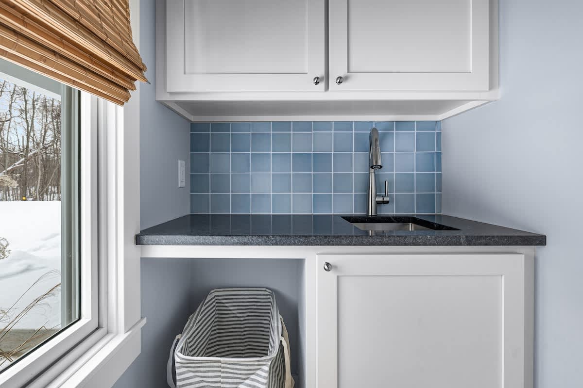 A bright laundry nook features light blue walls, white shaker-style cabinets, a dark countertop with an integrated sink, and a blue subway tile backsplash. A window with a bamboo blind offers a view of a snowy landscape.