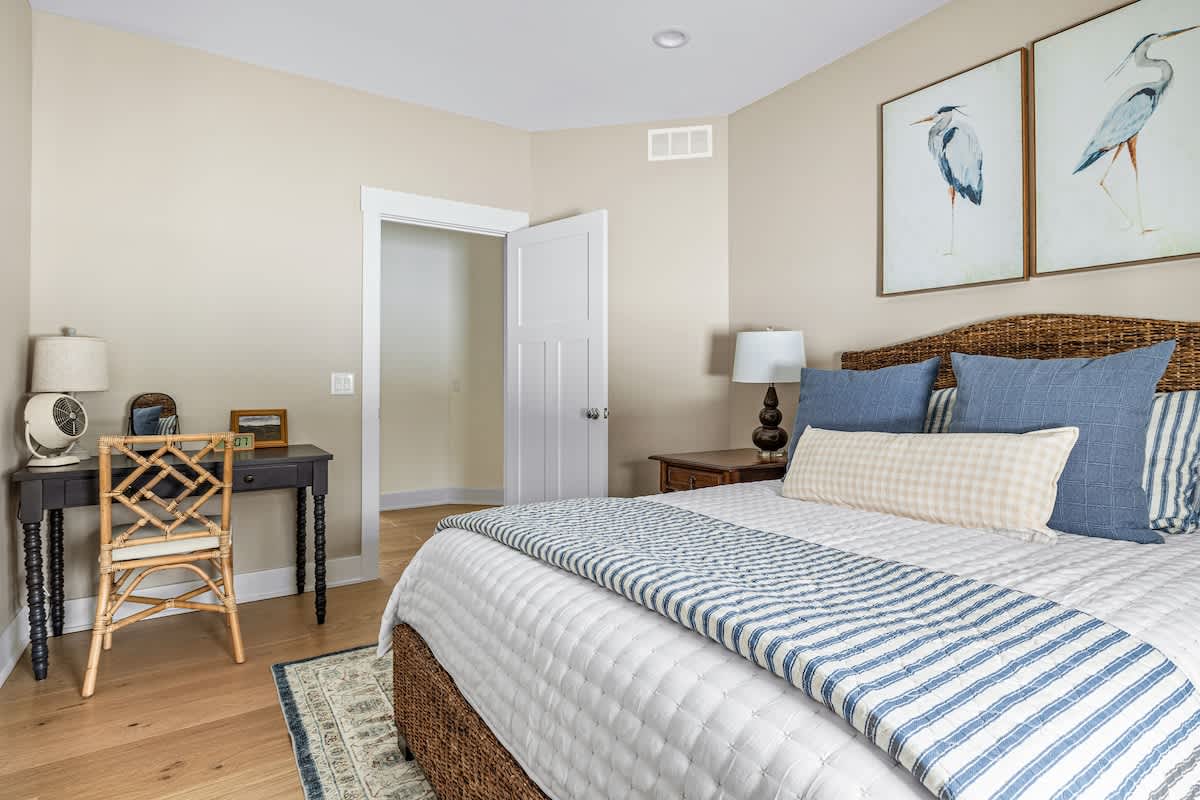 Another view of bedroom-2, highlighting a small dark wood desk with turned legs and a woven chair in the corner, next to a window with a bamboo blind. The wicker bed with blue and white bedding and heron artwork is visible, creating a serene and functional space.