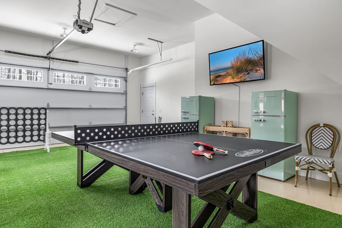A bright game room, set in what appears to be a garage with a large garage door, features a black ping pong table on a green artificial turf rug. Two retro-style mint green refrigerators and a wall-mounted TV are visible, along with a large connect-four game.
