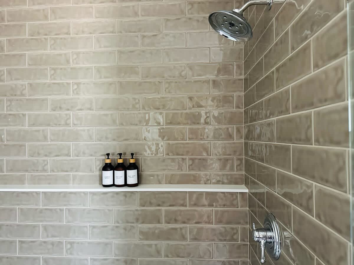 Modern shower with light gray subway tiles and a chrome shower head. A white shelf holds three dark-colored dispenser bottles for toiletries.