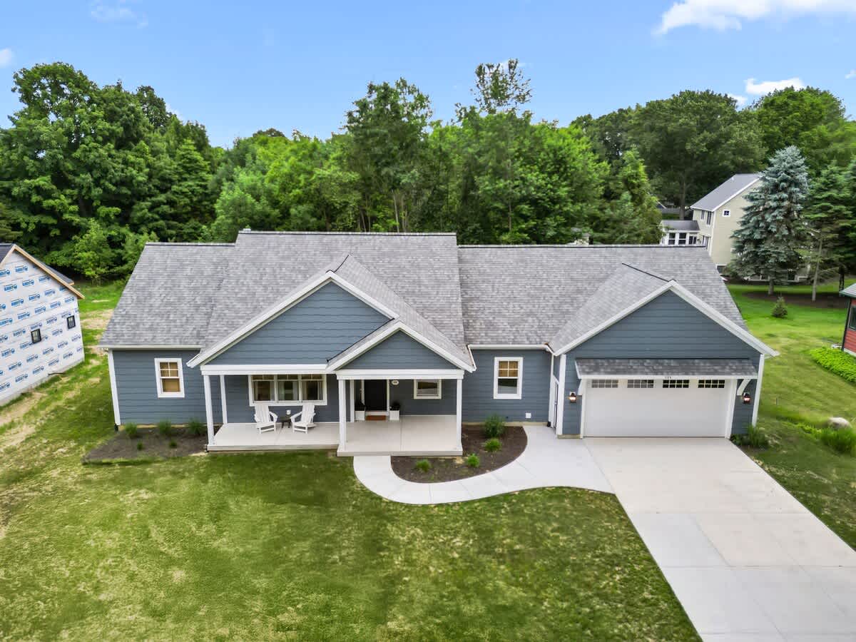 An aerial view showcases the blue-sided ranch home with a gray shingle roof, nestled among lush green trees. A winding concrete pathway leads to the covered front porch, and a large driveway connects to the two-car garage.