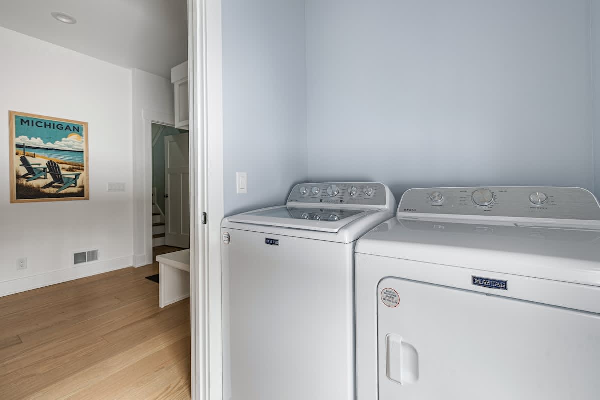 A dedicated laundry area features a white top-loading washing machine and a front-loading dryer, both by Maytag, set against light blue walls. Hardwood floors extend into an adjacent hallway with a framed Michigan poster.