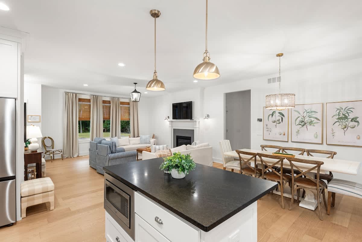 An open-concept living, dining, and kitchen area features light hardwood floors and white walls. The living room has a sofa and armchair, the dining area a long table, and the kitchen an island with pendant lights, all bathed in natural light.