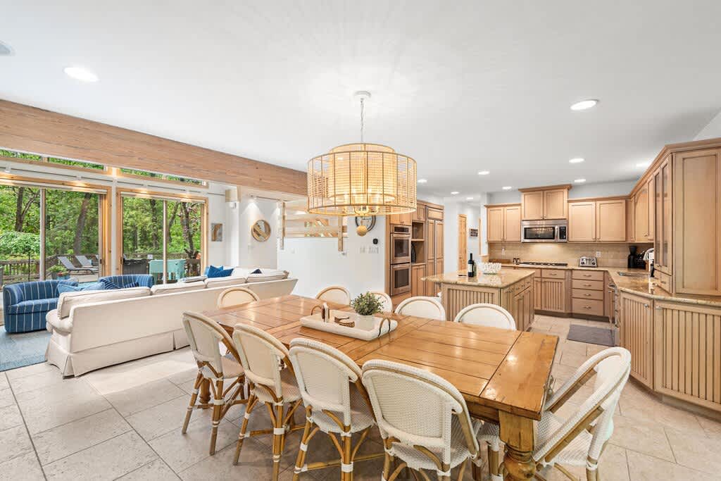Spacious open-concept dining area with a large wooden table and eight white wicker chairs, illuminated by a modern drum pendant light. The dining space flows into a well-equipped kitchen with light wood cabinetry and an island.