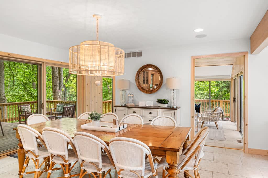 A bright dining room features a large wooden table with white wicker-back chairs, illuminated by a large circular woven pendant light, with sliding glass doors opening to a lush green outdoor deck.
