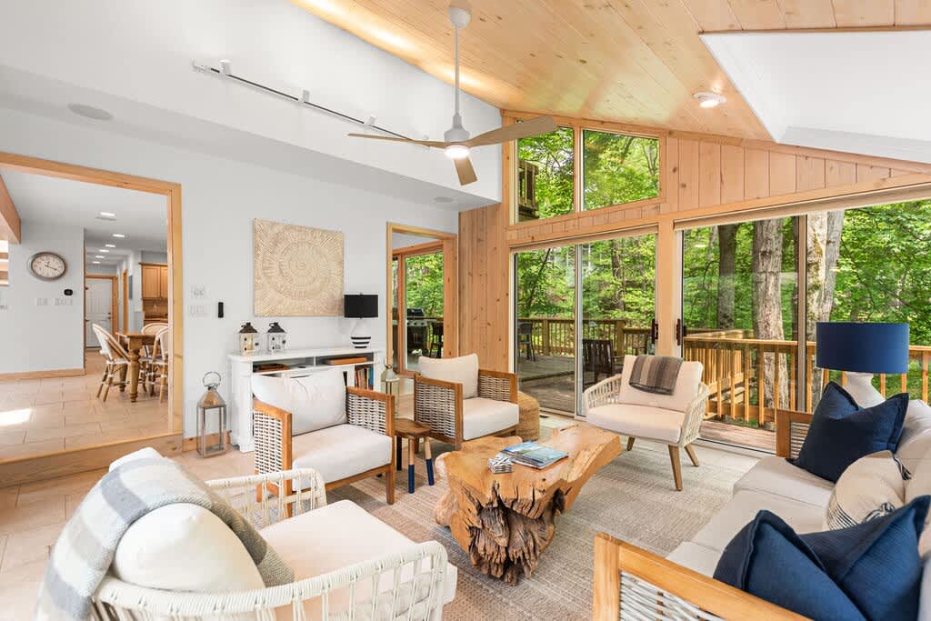 A bright and airy living room with a vaulted wooden ceiling and large windows, furnished with comfortable seating and a unique tree-trunk coffee table, offering views of the surrounding greenery and connecting to a screened porch.