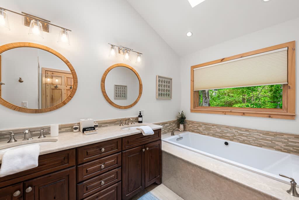 Bright bathroom showcasing a double vanity with dark wood cabinetry, a large soaking tub beneath a window, and a tiled backsplash, with a view of lush greenery outside.