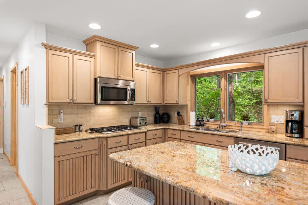 A well-lit kitchen area showcasing light wood cabinetry, speckled granite countertops, and a gas range with a microwave above, centered around a large window overlooking lush greenery.