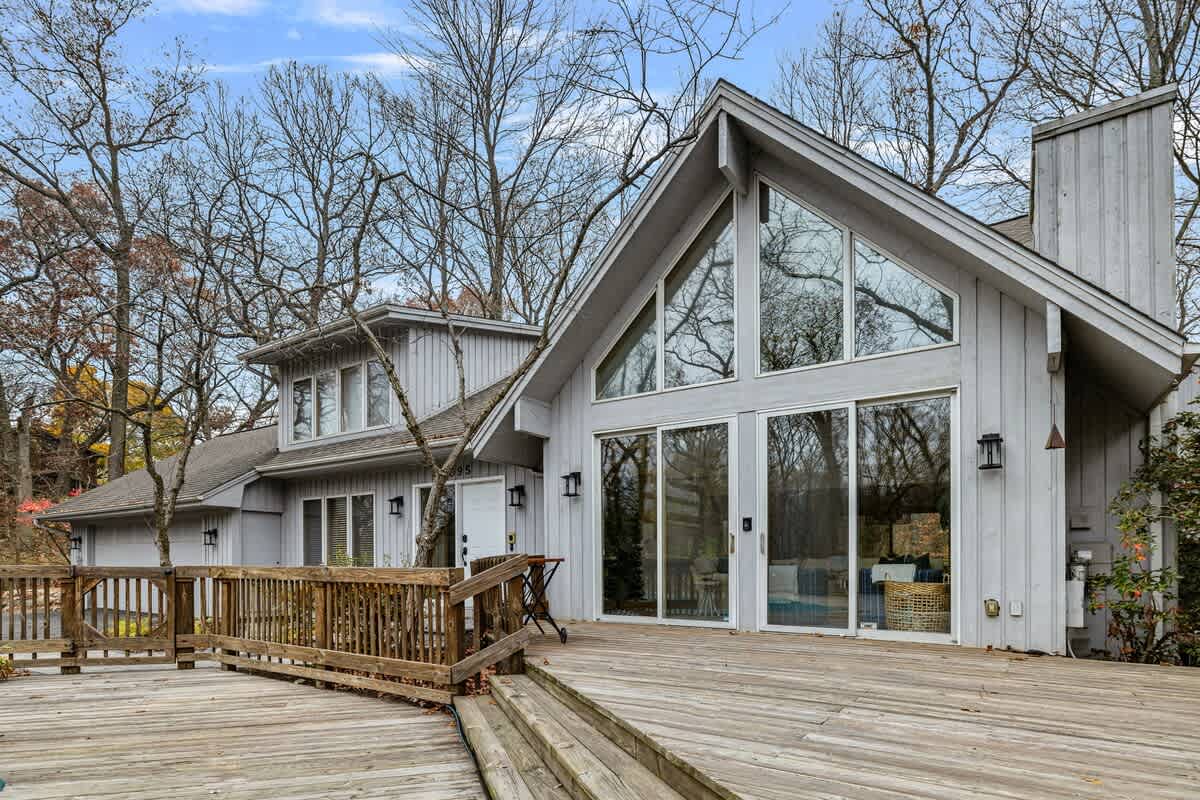 The back of a modern A-frame style house with light gray siding and large windows, featuring a spacious wooden deck that wraps around the structure, surrounded by tall, bare trees.