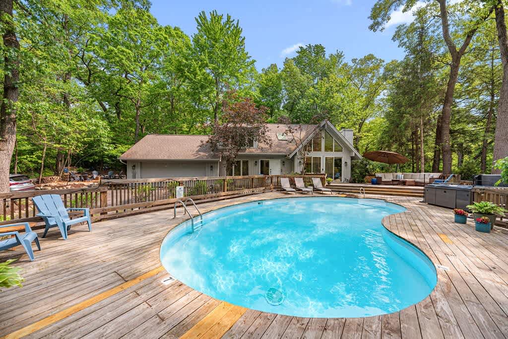 Aerial view of the property's rear, showcasing a large wooden deck with a kidney-shaped swimming pool and a hot tub, surrounded by mature green trees. The house features a light-colored siding and a prominent A-frame section with large windows.