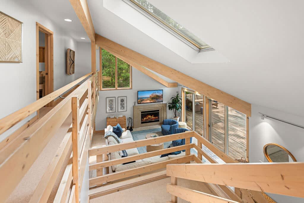 Elevated view of a bright living room featuring a vaulted ceiling with exposed wooden beams and skylights, and a large flat-screen TV above a fireplace. Comfortable sofas and armchairs are arranged on a rug, with large windows offering outdoor views.