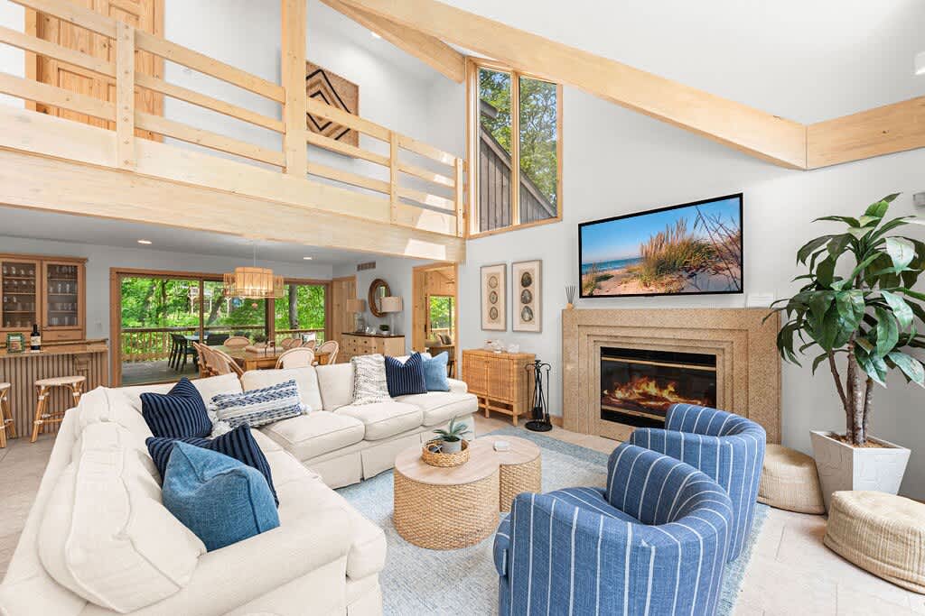 Open-concept living room with a cream sectional sofa and blue accent pillows, facing a stone fireplace with a mounted TV. The space features a vaulted ceiling, exposed wooden beams, and a view into the dining area and kitchen.